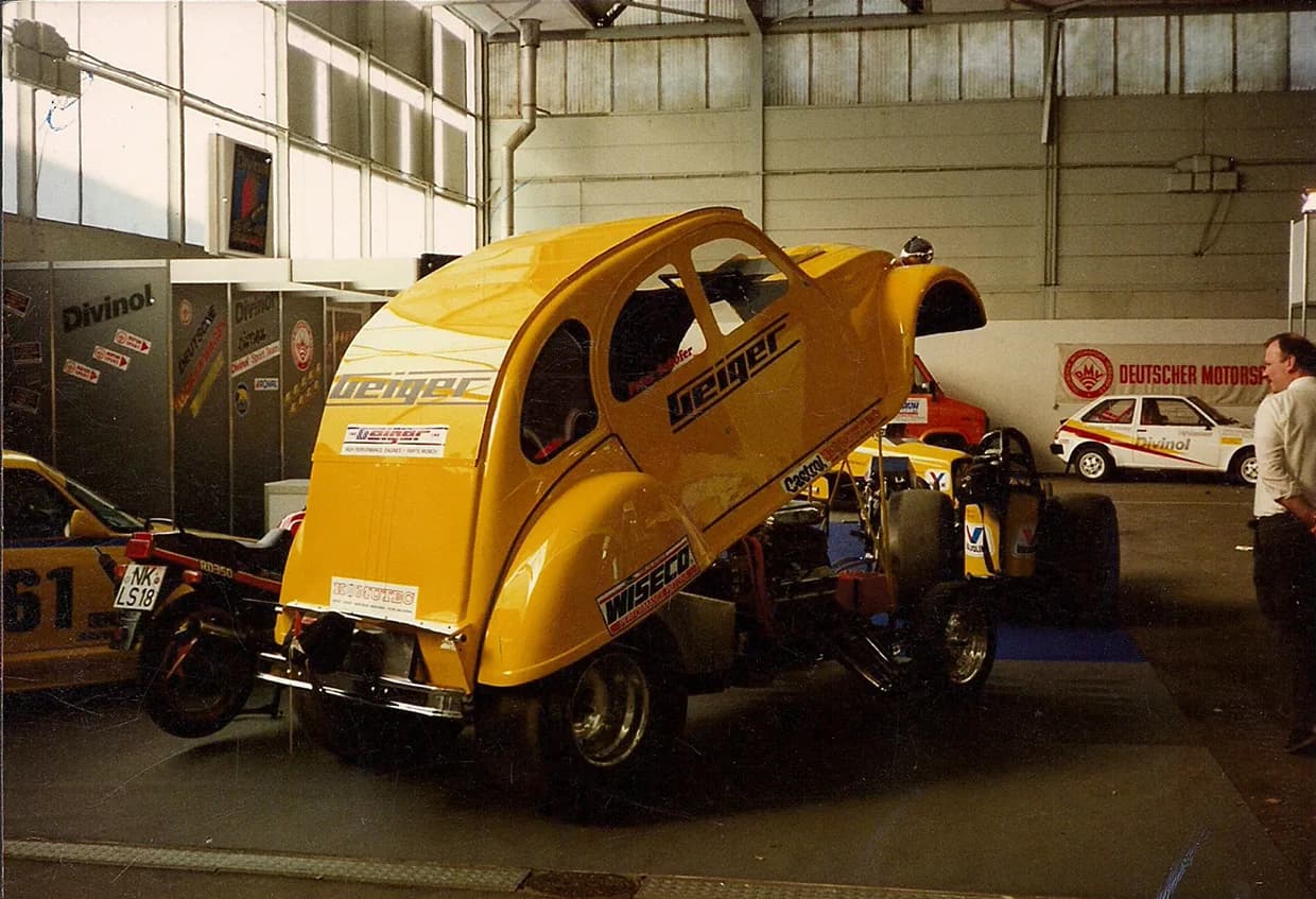 Gelbe Ente 2CV mit Geiger-Schriftzug und freigelegtem Motor auf einem Messestand in den 80er Jahren