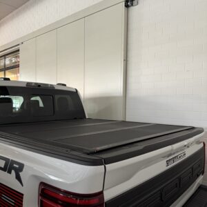 Rück- und Seitenansicht eines weißen Ford Raptor mit geschlossener schwarzer Ladeflächenabdeckung, geparkt in heller Garage.