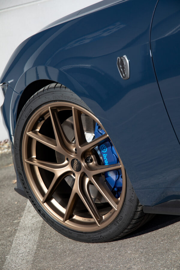 Close-up of the front left wheel of a dark blue Ford Mustang with a bronze multi-spoke rim, blue brake caliper, and "Dark Horse" emblem on the fender.