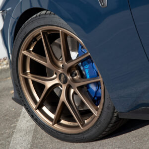 Nahaufnahme des vorderen linken Rads eines dunkelblauen Ford Mustang mit bronzefarbener Mehrspeichen-Felge, blauer Bremszange und „Dark Horse“-Emblem am Kotflügel.