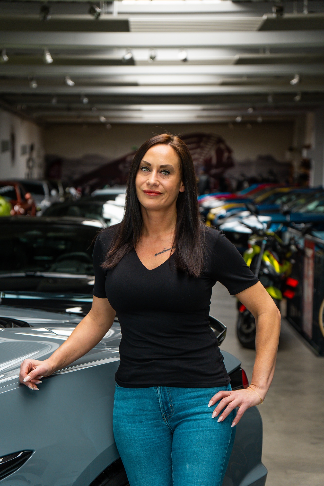 Frau mit dunklem Haar steht in einer exklusiven Fahrzeughalle neben einem grauen Sportwagen, im Hintergrund sind mehrere hochwertige Autos und Motorräder zu sehen.